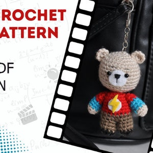 May include: Crochet pattern PDF in English for a small, beige teddy bear wearing a red shirt with a yellow lightning bolt and blue sleeves. The bear is attached to a silver keychain.