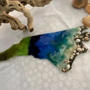 May include: A resin art piece in the shape of North Carolina, featuring a coastal design. The artwork incorporates shades of blue, green, and white, with embedded seashells and starfish. Driftwood and seashells are in the background.