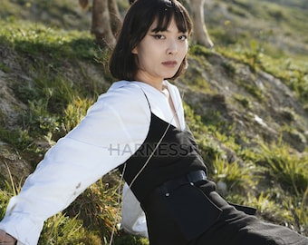 Puede incluir: Una mujer con una camisa blanca y un vestido negro se sienta en una ladera cubierta de hierba. Tiene el pelo negro corto y mira directamente a la cámara.