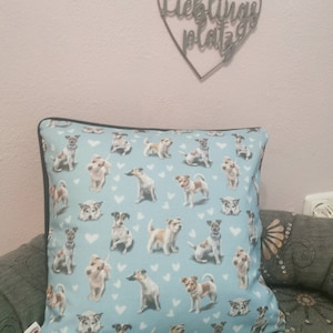 Könnte beinhalten: Ein blaues Kissen mit schwarzem Rand zeigt ein sich wiederholendes Muster aus weißen Herzen und Jack Russell Terriern. Das Kissen liegt auf einem Sofa mit Blumenmuster. Ein herzförmiges Schild aus Metall mit den deutschen Worten "Lieblings Platz" hängt an der Wand hinter dem Kissen.
