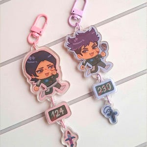 Könnte beinhalten: Zwei Acryl-Schlüsselanhänger mit Chibi-Figuren in dunkelgrünen Trainingsanzügen. Einer hat einen rosa Verschluss, die Nummer 124 und einen kleinen Schwertanhänger. Der andere hat einen helllila Verschluss, die Nummer 230 und ein kleines Kreuz.