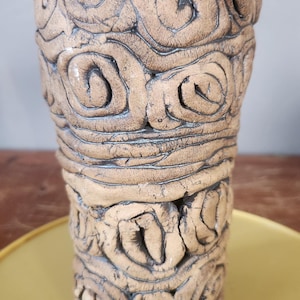 May include: A handcrafted, beige ceramic vase with a textured surface. The vase features a repeating pattern of swirling, raised designs. The vessel is cylindrical with a slightly flared top, and sits on a yellow surface.