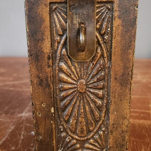 May include: A rectangular, antique-style box with a hinged lid and clasp. The box is made of a dark, aged metal with a decorative sunburst design on the front. The clasp is a darker, contrasting metal.