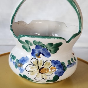 May include: A white ceramic basket with a handle, adorned with hand-painted blue and yellow floral designs and green accents. The rim and handle are trimmed with green paint. A decorative piece for the home.