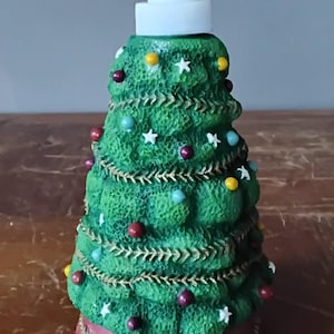 May include: A Christmas tree-shaped soap dispenser. The green dispenser is decorated with colorful ornaments, white star accents, and a gold-colored garland. The base is red with a decorative pattern, and the pump is white.