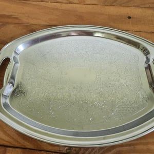 May include: An oval silver-toned metal serving tray featuring a polished smooth rim and integrated handles on both sides. The central surface displays an intricate, embossed floral and scrollwork pattern with a blank oval space in the center.