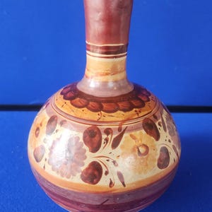 May include: A round ceramic vase with a narrow neck, featuring a cream-colored base decorated with hand-painted brown and reddish-purple floral patterns, scalloped border details, and horizontal bands encircling the neck and the lower body.