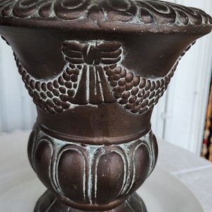 May include: A brown ceramic planter with a decorative design of a winged figure and a grapevine pattern. The planter has a distressed finish with a light green patina.