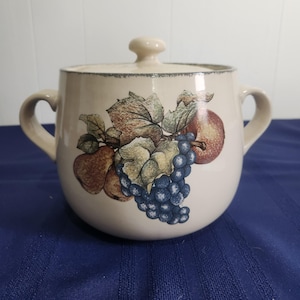May include: Cream-colored ceramic casserole dish with a lid and two handles. The dish is decorated with a colorful illustration of grapes, apples, and leaves. The lid has a small knob for easy handling. The dish is on a blue surface.