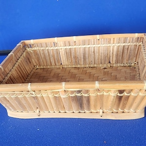 May include: Rectangular woven bamboo basket with a natural wood finish. The basket features a woven base and sides constructed from vertical bamboo pieces, secured with thin, light-colored twine. The basket's design is simple and functional, suitable for storage or display.