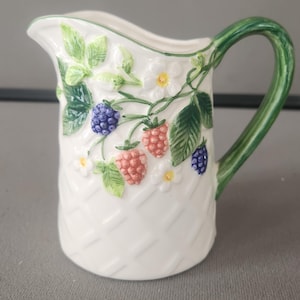 May include: White ceramic pitcher with a green handle and rim. The pitcher is decorated with raised relief images of berries, leaves, and flowers. The handle is a solid green color, and the pitcher has a basket weave pattern.