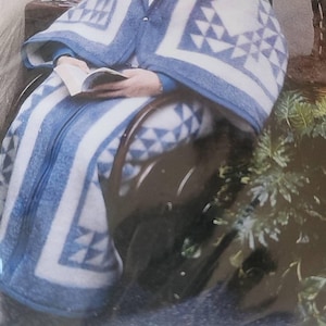 May include: A blue and white patterned blanket, measuring 55 x 67 inches, is draped over a person. The blanket features a geometric design and is labeled "Shaker blue". The person is seated in a chair and holding a book.