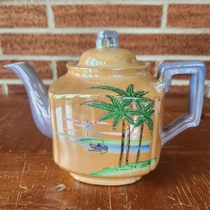 May include: A vintage teapot with a pastel purple handle and a light brown body. The teapot has a scene of palm trees and a boat on a lake painted on the front.