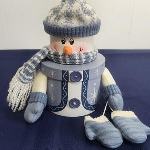 May include: A decorative snowman figurine crafted from a cylindrical box, featuring a blue and white knit hat, a matching striped scarf, and two dark blue buttons on the front. The snowman has soft arms with attached blue mittens and two extra mittens.