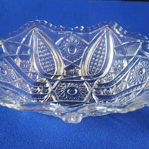 May include: A clear pressed glass bowl featuring a complex geometric pattern of starbursts and elongated oval motifs. A yellow measuring tape is positioned along the side, indicating the bowl has a width of approximately 6 inches at its widest point.