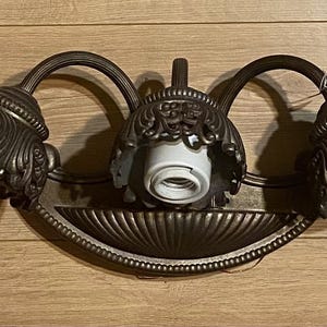 May include: A bronze-colored three-light fixture with ornate, decorative shades. The fixture has a curved design and is suitable for a bathroom or vanity.