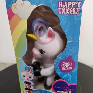 May include: A boxed toy featuring a white unicorn with a pink and blue mane, a yellow horn, and black hooves. The box is blue with the words "HAPPY UNICORN" and "40+ Sounds". The text "I react to touch, motion & sounds" is also visible.