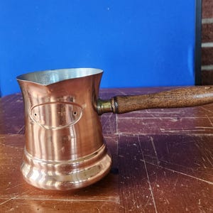 May include: A copper coffee pot with a wooden handle. The pot has a shiny, polished finish and a small spout. The handle is attached with a brass fitting. The pot is designed for stovetop use.