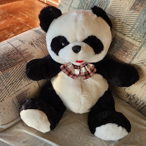 May include: A black and white plush panda bear wearing a red and white plaid bow tie. The panda is sitting on a patterned fabric surface.