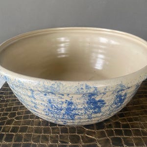 May include: A large, off-white ceramic bowl with a wide rim and a blue speckled pattern. The bowl has a smooth interior and a slightly textured exterior. This vintage-style bowl is suitable for kitchen use.