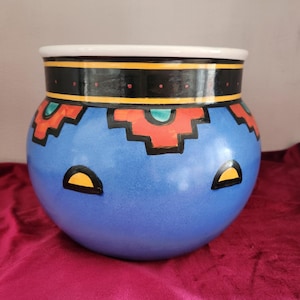 May include: A blue ceramic planter with a geometric pattern in red, yellow, and turquoise. The planter has a white rim with a black and yellow band.