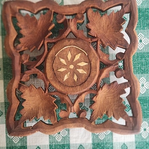May include: A square, wooden trivet with a carved floral design. The trivet features a central circular element with a starburst pattern, surrounded by carved leaves and vines. The wood has a warm, brown tone.