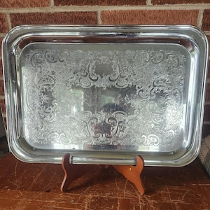 May include: A rectangular silver-toned serving tray featuring a polished metallic finish and an intricate, embossed scrollwork pattern across the central surface. The tray has a smooth, raised border and is displayed on a brown wooden stand.