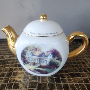 May include: White ceramic teapot with gold accents on the spout, handle, and lid. The teapot features a painted scene of a house and the text "Home Is Where The Heart Is".