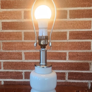 May include: A white, stacked-base table lamp with a silver metal base and accents. The lamp has a bright, round light bulb and a silver metal shade holder. The background is a brick wall and a framed picture.