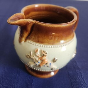 May include: A ceramic creamer with a brown rim and handle, and a light green body. The body features a decorative relief of a person riding a horse. The creamer has a spout for pouring.