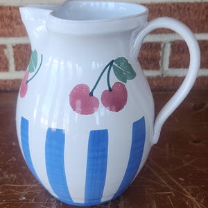 May include: White ceramic pitcher with a handle and spout. The pitcher is decorated with blue vertical stripes and pink cherry designs with green leaves. The pitcher is likely used for serving beverages.