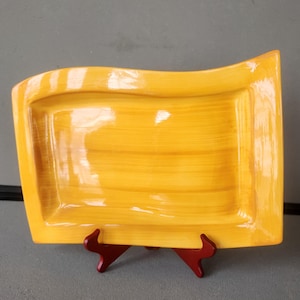 May include: A rectangular, yellow ceramic serving tray with a wavy edge. The tray is supported by a red stand. The tray has a glossy finish and a brushed texture.