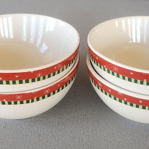 May include: Two stacks of white ceramic bowls with a red band featuring green stripes and white snowflake designs. The bowls are stacked on a gray surface.