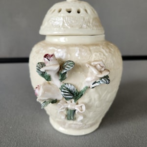 May include: Cream-colored ceramic jar with a lid, adorned with embossed floral patterns and three-dimensional rose accents in white, pink, and green. The lid features small ventilation holes and a decorative knob.