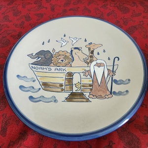 May include: A round ceramic plate with a blue rim and a beige center. The plate features a colorful illustration of Noah's Ark with various animals, including an elephant, lion, giraffe, and dove. The words "Noah's Ark" are written on the ark.