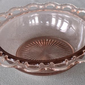 May include: A clear, pink-tinted glass bowl with a scalloped, openwork rim. The bowl has a circular, textured base with a sunburst pattern. The rim and base have a darker pink hue, creating a contrast with the clear sides.