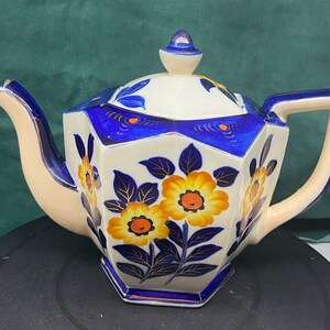 May include: A white teapot with a blue and gold floral design. The teapot has a rounded body and a spout on the side. The handle is on the top of the teapot.