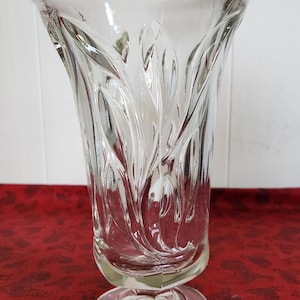 May include: A clear glass vase with a flared, scalloped rim and a pedestal base. The vase features a repeating leaf-like pattern on its sides, creating a textured appearance. The vase is empty and sits on a red surface.