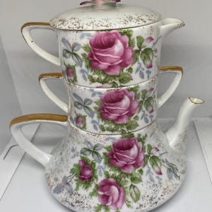 May include: A three-tiered porcelain teapot with a floral design. The teapot is decorated with pink roses and green leaves on a white background. The teapot has a gold rim and a lid with a pink rose on top.