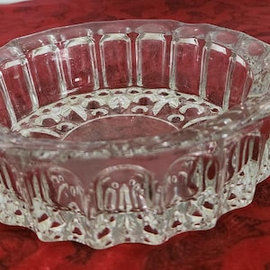 May include: A clear, oval-shaped glass ashtray with a decorative, scalloped rim. The ashtray features a detailed, embossed design around the sides and base. The glass has a textured appearance, and the ashtray sits on a red surface.