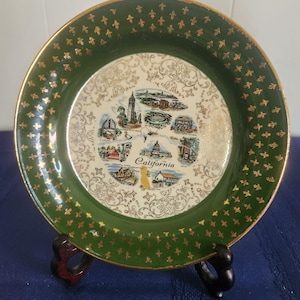 May include: A round decorative plate featuring a dark green border with a gold fleur-de-lis pattern and a gold rim. The center displays a white background with gold filigree and a collage of California landmarks with the text California.
