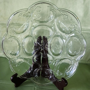 May include: Clear glass egg serving platter with floral and leaf detailing. The platter has 12 egg-shaped indentations and is supported by a dark brown wooden stand. The platter is on a green surface.
