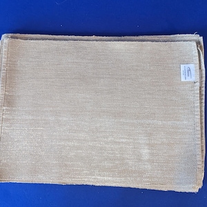 May include: A stack of beige rectangular placemats with a woven texture. The placemats have a subtle sheen and a label in the corner that reads "AMAN". The placemats are neatly folded and appear to be made of a durable material.