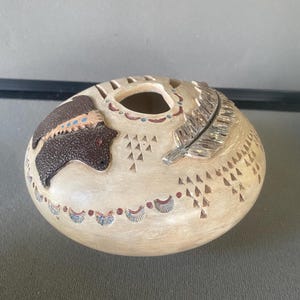 May include: A round, cream-colored ceramic vessel with a textured surface. It features a raised dark brown bear figure, a carved feather detail, and small triangular patterns. The rim is decorated with a series of small blue and red painted dots.