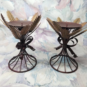 May include: Two metal candle holders with a brown finish and gold accents. Each holder features a design of wheat stalks with a bow at the base.