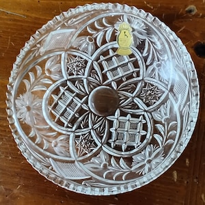 May include: Clear glass bowl with intricate floral and geometric cut patterns. The bowl features a scalloped edge and a central, circular design. A gold-colored sticker is affixed to the bowl.