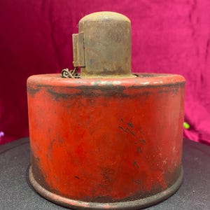 May include: A red metal container with a rusty metal lid and a chain attached. The container has a worn, vintage look.