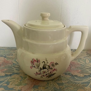 May include: Cream-colored ceramic teapot with a lid and handle. The teapot features a floral design with pink and white flowers. The teapot has a spout and a handle for easy pouring. The teapot is designed for brewing tea.