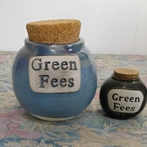 May include: Two ceramic jars with cork stoppers. The larger jar is blue with the words "Green Fees" on a white label. The smaller jar is black with the same label. Both jars are on a floral patterned surface.