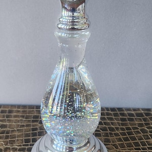 May include: A silver-colored candle holder with a clear, glitter-filled glass center. The base and top are silver, with a decorative, swirling design in the middle. The candle holder sits on a textured, brown surface.